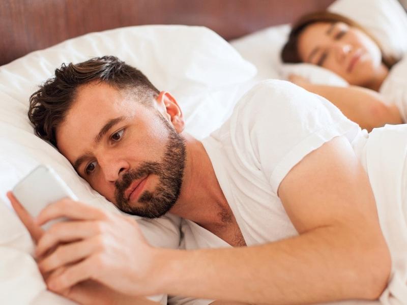 Hombre revisando su celular mientras su esposa duerme.