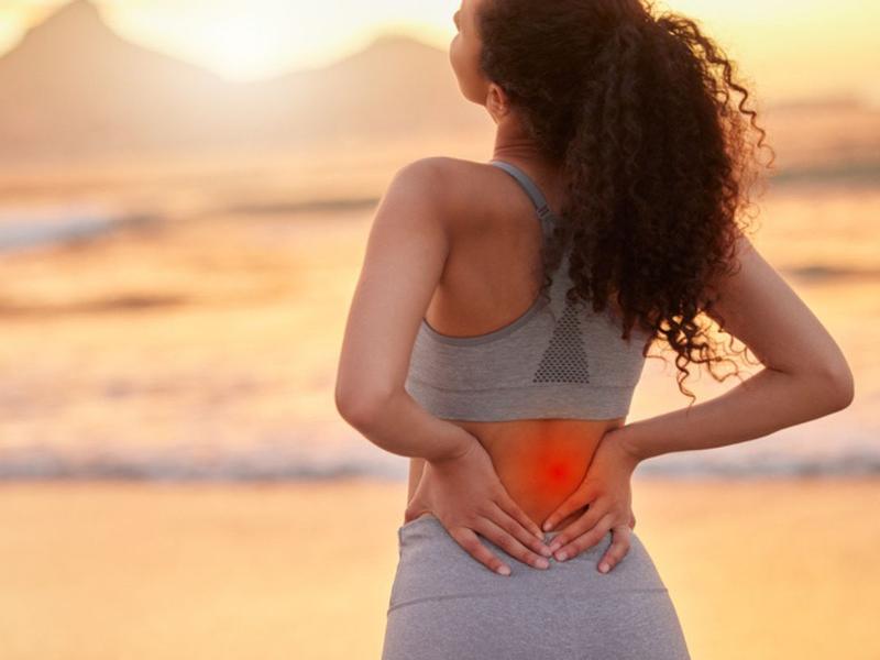 Mujer en la playa se toca la espalda porque no sabe los tips para un verano sin dolor de espalda
