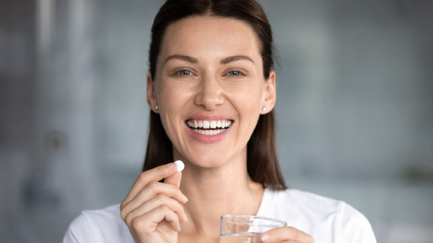 Mujer sonriente sosteniendo una pastilla en una mano y en la otra mano un vaso con agua.
