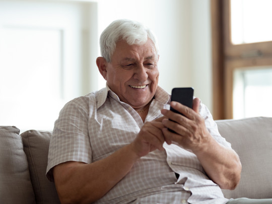 Hombre mayor sonriendo mientras mira en celular que Adultos mayores que usan internet tienen menor riesgo de demencia