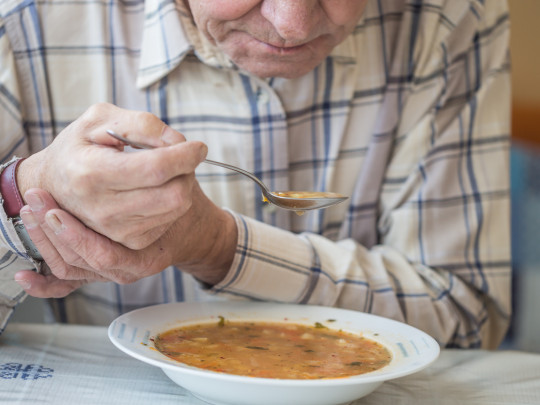 Hombre a punto de comer sopa se sostiene con ambas manos porque sabe que la soledad puede provocar Parkinson, revela estudio