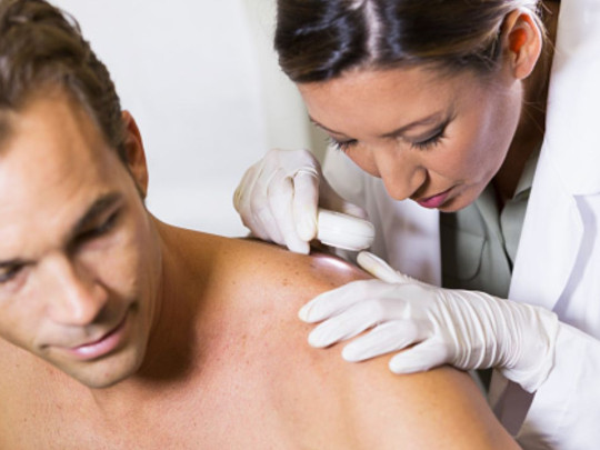 Dermatóloga examinando espalda de hombre para explicarle los 5 signos menos comunes del cáncer de piel